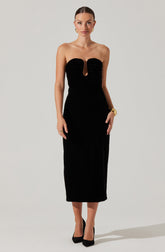 A woman is wearing a strapless, black, floor-length gown with a keyhole cutout in the center of the bodice. The top of the gown features gathered fabric around the bust area. She accessorizes with gold earrings, a gold bracelet, and rings. She is wearing black high heels.